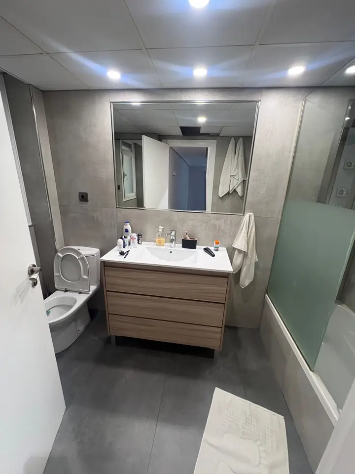 Foto real de un baño antes de la reforma: lavabo con mueble de madera, inodoro, bañera con mampara y azulejo gris
